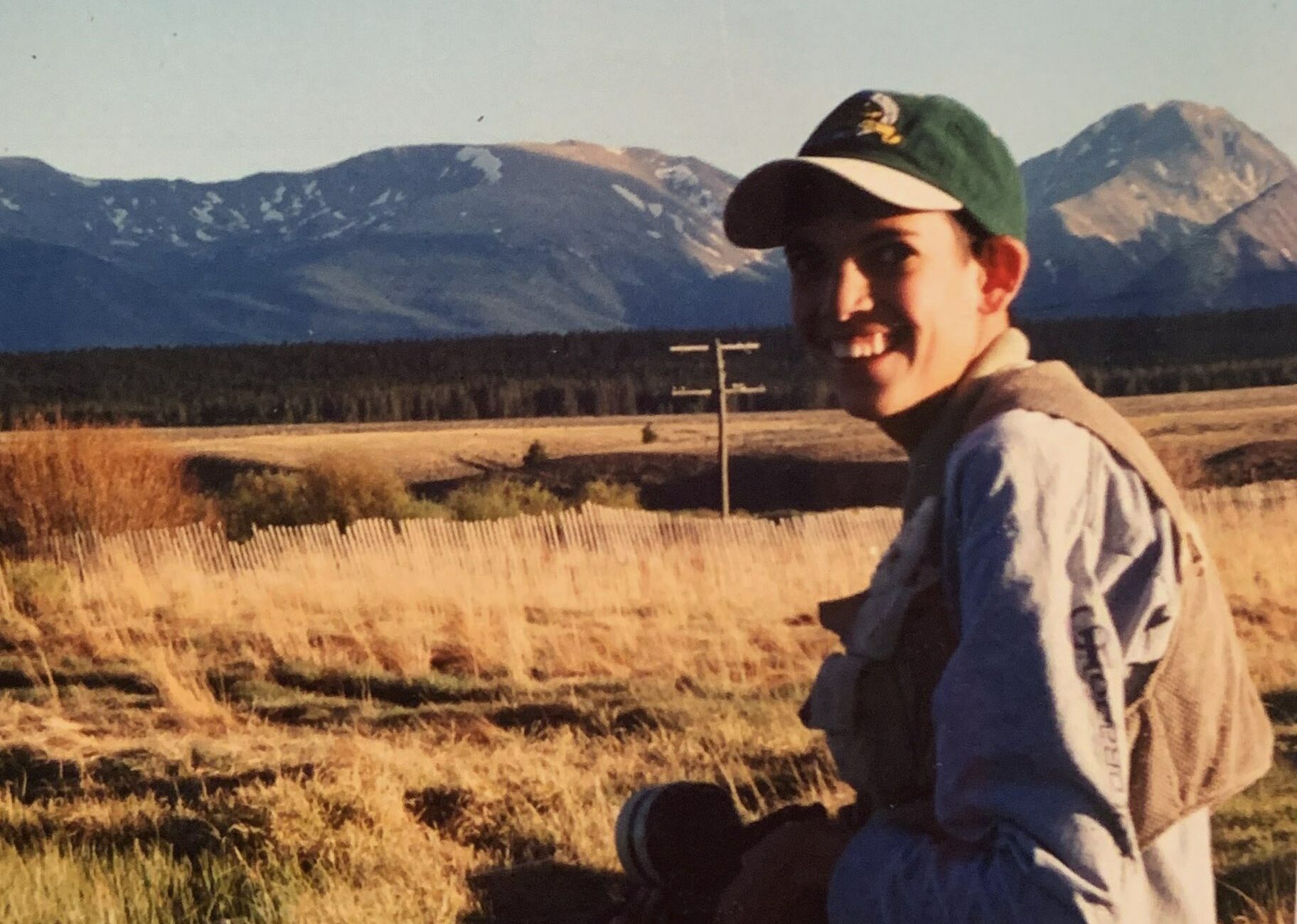 Geoffrey, a lanky adolescent with a baseball cap on his head, looks back at the camera, smiling broadly. He stands in a high mountain desert landscape with mountains rising up in the distance.