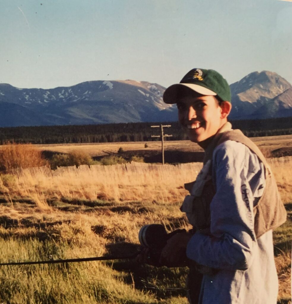 Geoffrey, a lanky adolescent with a baseball cap on his head, looks back at the camera, smiling broadly. He stands in a high mountain desert landscape with mountains rising up in the distance.